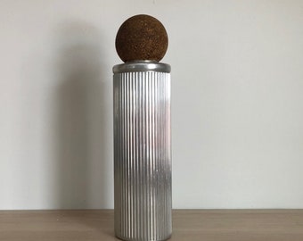 May include: A silver-colored, vertically-ribbed bottle with a cork ball stopper. The bottle has a cylindrical shape and sits on a light-colored wooden surface against a white wall. The cork ball is a dark brown color.