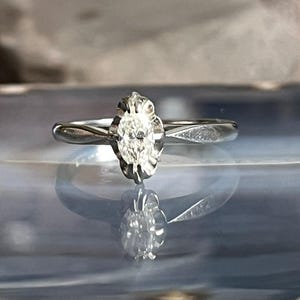 Puede incluir: Un anillo de compromiso de plata con un solo diamante claro de forma ovalada engastado en un engaste de forma floral.