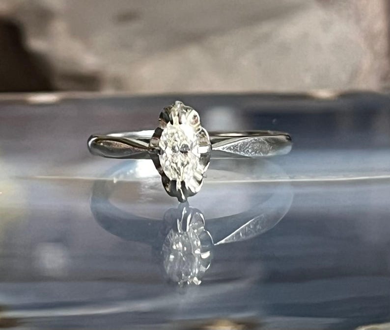Puede incluir: Un anillo de compromiso de plata con un solo diamante claro de forma ovalada engastado en un engaste de forma floral.