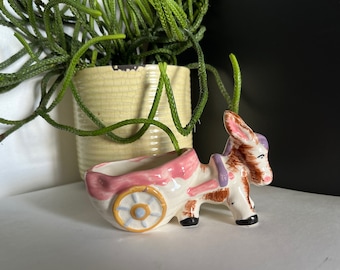 May include: A ceramic donkey planter with a pink and white wagon. The donkey is brown and white with purple accents. The wagon has a single yellow wheel with a blue center.