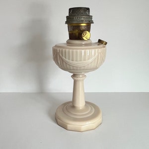 May include: A pale pink oil lamp with a fluted design and a decorative trim around the top. The lamp has a metal burner and a glass chimney.