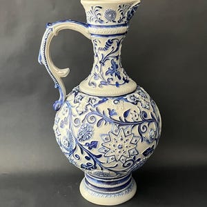 May include: A white and blue ceramic pitcher with intricate floral and star patterns. The pitcher has a rounded body, a handle, and a spout. The design features raised relief details and a decorative base.