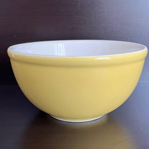 Peut inclure: Un saladier en céramique jaune avec un intérieur blanc. Le bol a une forme arrondie et une finition lisse et brillante. Il est posé sur une surface sombre.