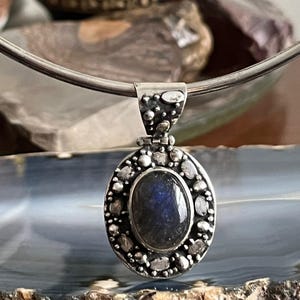 May include: A silver pendant with a dark blue labradorite gemstone set in a silver bezel. The pendant is surrounded by a silver halo of small beads and stones.