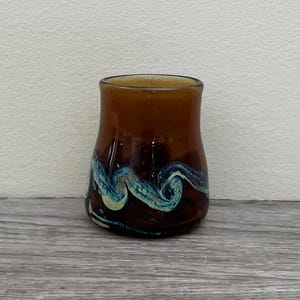 Peut inclure: Un gobelet en verre marron avec un motif ondulé bleu et blanc autour de la base. Le verre a une forme lisse et arrondie et un dessus légèrement évasé. Le gobelet est posé sur une surface en bois.