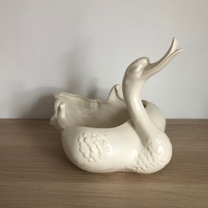 May include: Cream-coloured ceramic swan planter. The swan has a long, curved neck and a detailed body with textured feathers. The planter has a large opening for plants or decorative items. The swan is approximately 30 cm long.