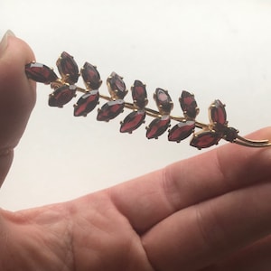 May include: A gold-tone brooch in the shape of a branch with maroon-colored glass stones.