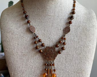 May include: A brown beaded necklace with a pendant and dangling amber-colored beads. The necklace features a detailed, ornate pendant and smaller decorative elements. The beads are various shapes and sizes, creating a layered effect. The necklace is displayed on a mannequin.