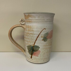 May include: A tall, handmade ceramic mug with a large handle and a decorative swirl at the top. The mug is off-white with brown speckles and features a painted floral design in pink and green.