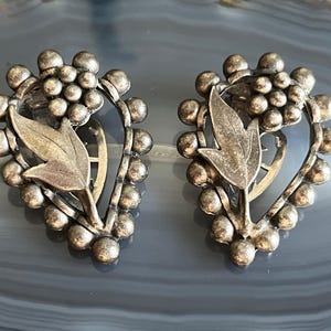 May include: A pair of silver tone earrings with a floral design. The earrings are shaped like a heart and have a cluster of small beads around the edge.