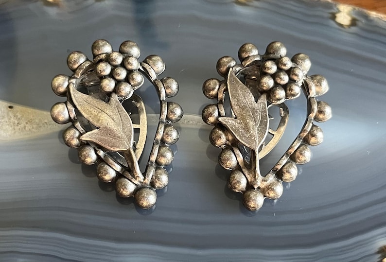 May include: A pair of silver tone earrings with a floral design. The earrings are shaped like a heart and have a cluster of small beads around the edge.