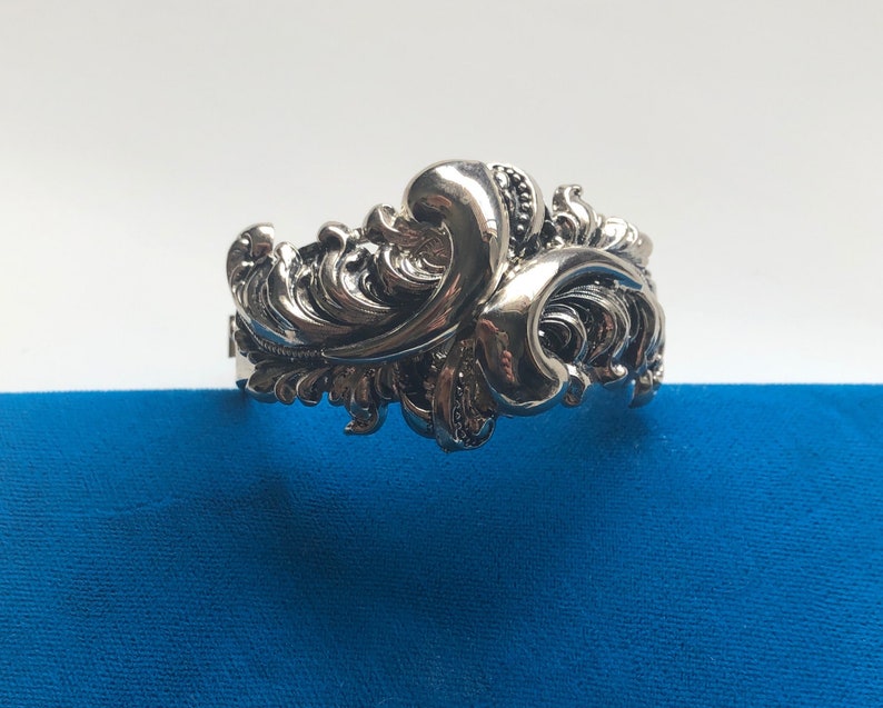 K&ouml;nnte beinhalten: Ein silberfarbenes, verziertes Armband mit floralem Design. Das Armband ist aus Metall und hat ein detailliertes, kompliziertes Muster.