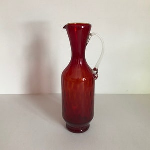 Może przedstawiać: Czerwony dzbanek szklany z przezroczystym uchwytem. Dzbanek ma żebrowany wzór i zaokrągloną podstawę.