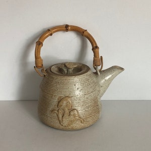May include: A ceramic teapot with a speckled glaze and a bamboo handle. The teapot has a raised design of a stylized figure on the side.