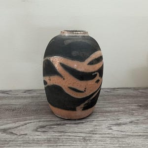 May include: A ceramic vase with a rounded body and a narrow neck. The vase has a black background with a swirling, abstract design in a light orange colour. The vase is approximately 15 cm tall.
