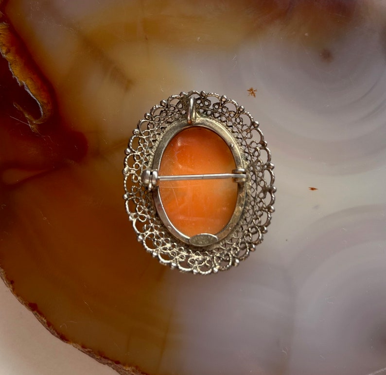 May include: A cameo brooch with a delicate silver filigree design. The cameo features a carved portrait in shades of orange and white.