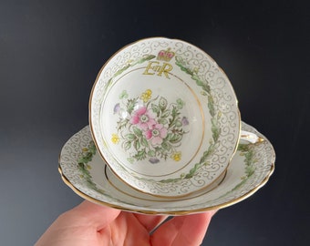 Könnte beinhalten: Eine weiße Teetasse und Untertasse mit goldfarbenem Rand und Blumenmuster. Die Teetasse zeigt eine Krone und die Buchstaben "ER" in Gold.