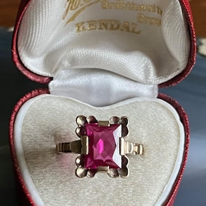 May include: A red velvet ring box with a white satin interior. The box is open and a gold ring with a large, square, ruby-red stone is inside. The box has the text "Watchman & Jewell H. Hoggan Bros. Goldsmith Kendal" on the inside lid.