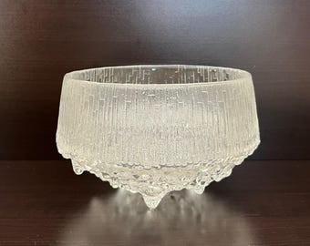May include: A clear, textured glass bowl with a wide, cylindrical shape. The bowl features a vertical, ridged pattern on the sides and a textured, droplet-like base. The bowl is designed to sit on three small feet.