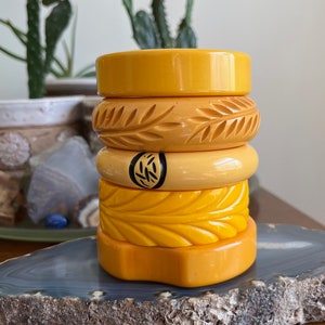 Puede incluir: Una pila de cinco pulseras de plástico amarillo con diferentes diseños tallados. Las pulseras están apiladas sobre una rebanada de ágata gris y blanca.