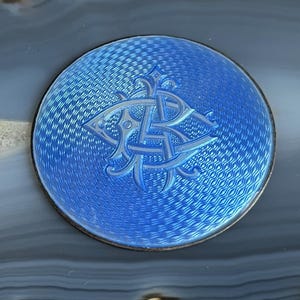 May include: A round, blue enamel object with a silver border. The blue surface has a textured pattern and features a light blue monogram design. The object is set against a gray and white background.