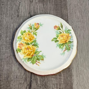 Puede incluir: Bandeja decorativa redonda blanca con borde festoneado y borde dorado. La bandeja presenta un diseño floral de rosas amarillas y hojas verdes. La bandeja tiene aproximadamente 20 cm de diámetro.