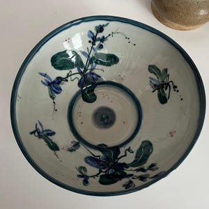 May include: A white ceramic bowl with a dark teal rim and a central dip. The bowl is decorated with blue and green floral patterns. The bowl has a diameter of approximately 12 inches.