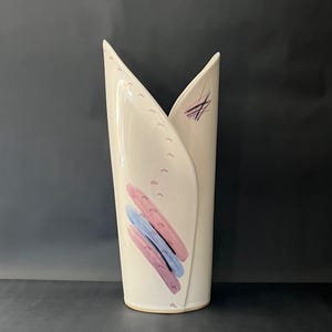 Peut inclure: Un vase en céramique blanc cassé de forme pliée unique. Il est orné de coups de pinceau abstraits roses, bleus et violets. De petits points roses et des lignes violettes ornent la surface. Le vase présente une esthétique moderne et minimaliste.