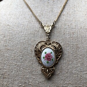 May include: A gold chain necklace with a heart-shaped pendant featuring a white oval cameo with a pink rose and green leaves.