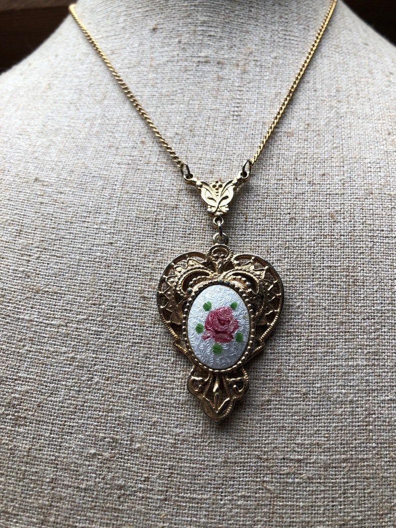 May include: A gold chain necklace with a heart-shaped pendant featuring a white oval cameo with a pink rose and green leaves.