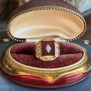 May include: A gold ring featuring a square, deep red stone and a diamond-shaped detail. The ring is presented in an open, vintage jewellery box with a red velvet interior and a gold-edged exterior.