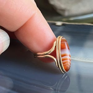 以下が含まれることがあります： 赤と白の瑪瑙の石が付いた金の指輪。石は楕円形で、縞模様があります。