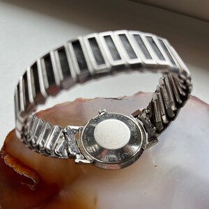 May include: A silver wristwatch with a round face and a silver metal band. The back of the watch has the text "DUSTPROOF" engraved on it.