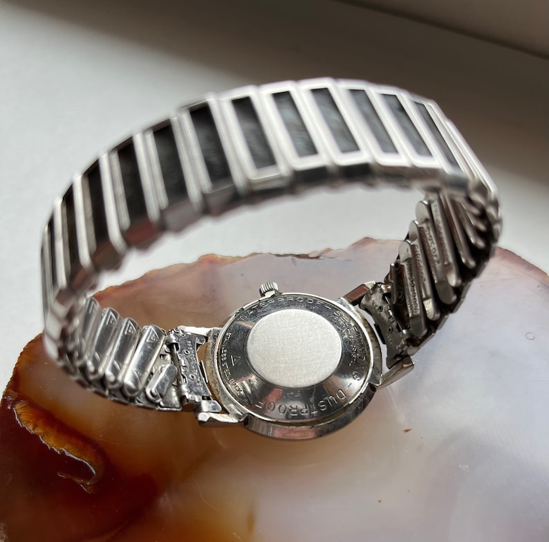 May include: A silver wristwatch with a round face and a silver metal band. The back of the watch has the text "DUSTPROOF" engraved on it.