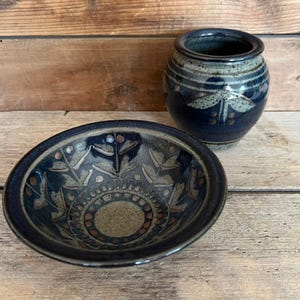 May include: A dark blue ceramic bowl and vase set. The bowl features a circular design with leaf and dot patterns. The vase is cylindrical with a dragonfly design. Both pieces have a rustic, handmade appearance.