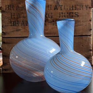 May include: Two glass vases with a light blue background and swirling stripes of brown and blue. The vases have a bulbous base and a narrow neck. The background is a wooden crate with faded text.