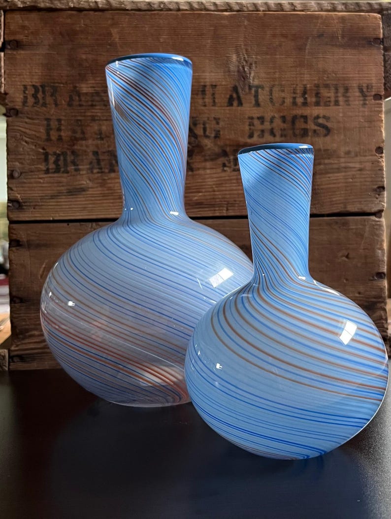 May include: Two glass vases with a light blue background and swirling stripes of brown and blue. The vases have a bulbous base and a narrow neck. The background is a wooden crate with faded text.