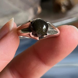 May include: A silver ring featuring a dark, rounded gemstone. The ring's band is a polished silver, and the gemstone is a deep, possibly iridescent, color. The ring is held between fingers, showcasing its design and details.