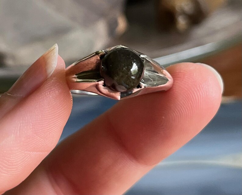 May include: A silver ring featuring a dark, rounded gemstone. The ring's band is a polished silver, and the gemstone is a deep, possibly iridescent, color. The ring is held between fingers, showcasing its design and details.
