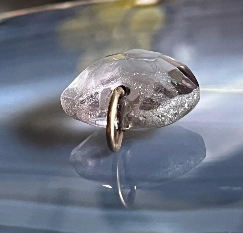 May include: A clear, faceted crystal pendant with a gold-colored metal loop. The crystal has a rounded shape with multiple facets, reflecting light. The pendant is suspended by a small, circular loop.