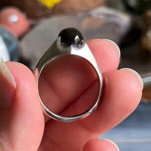 May include: A silver ring with a large, dark, round stone. The ring has a simple, modern design with a wide band and a bezel setting for the stone. The stone appears to be polished and reflective.