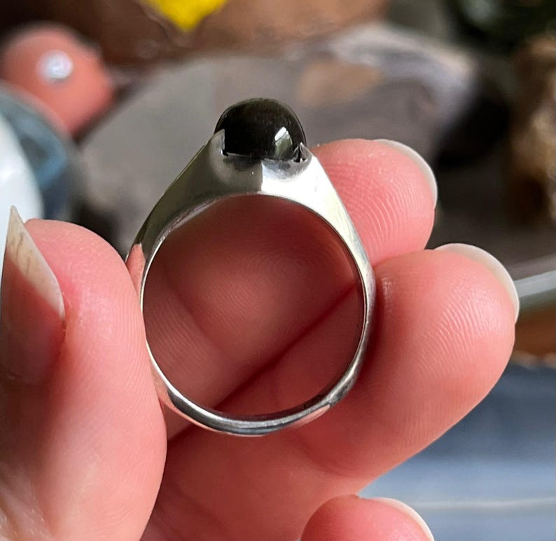 May include: A silver ring with a large, dark, round stone. The ring has a simple, modern design with a wide band and a bezel setting for the stone. The stone appears to be polished and reflective.