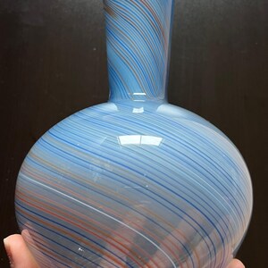 May include: A blue glass vase with a round base and a slender neck. The vase features a striped pattern in shades of blue, white, and orange. The vase is held in a hand.