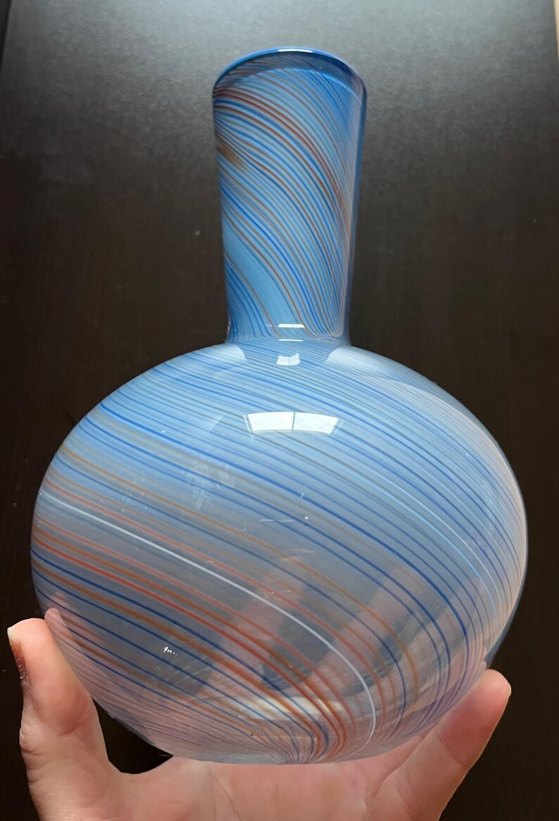 May include: A blue glass vase with a round base and a slender neck. The vase features a striped pattern in shades of blue, white, and orange. The vase is held in a hand.