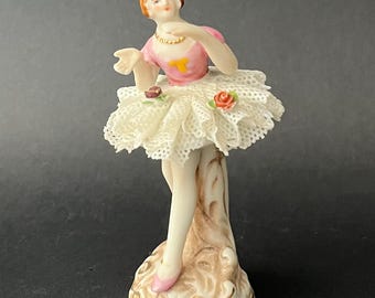 May include: A porcelain figurine of a ballerina in a pink top and white lace tutu. The figurine has a gold necklace and a small yellow accent on the bodice. The base is decorated with gold accents and the figurine is approximately 6 inches tall.