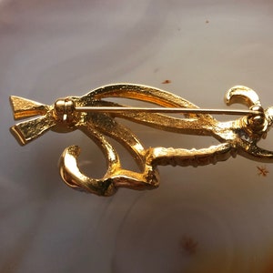 May include: Gold-toned brooch with a stylized design featuring a bird with a long tail. The brooch has a pin and catch closure on the back.