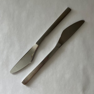 May include: Two stainless steel knives with a simple, modern design. The blades are smooth and slightly curved, with a polished finish. The handles are rectangular and also have a polished finish. The knives are set against a white background.
