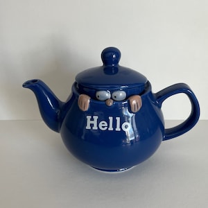 May include: A blue ceramic teapot with a lid and a handle. The teapot has a white inscription that says "Hello" and two eyes peeking out from under the lid.