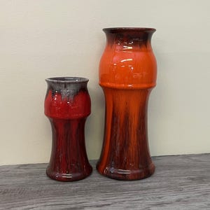 Puede incluir: Dos jarrones de cerámica con acabado brillante. El jarrón más grande es naranja y marrón, mientras que el más pequeño es rojo y marrón. Ambos jarrones tienen una forma única y acampanada y son adecuados para la decoración del hogar.