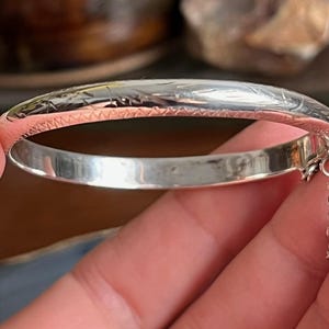 May include: A silver bangle bracelet with etched floral patterns and a small chain. The bracelet has a polished finish and a simple, elegant design. The chain is attached to the bracelet with a small loop.